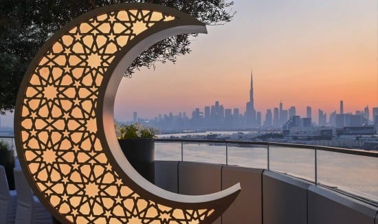 Su una terrazza una enorme mezzaluna illuminata, tipica decorazione del Ramadan, che abbraccia da lontano lo skyline di Dubai al tramonto